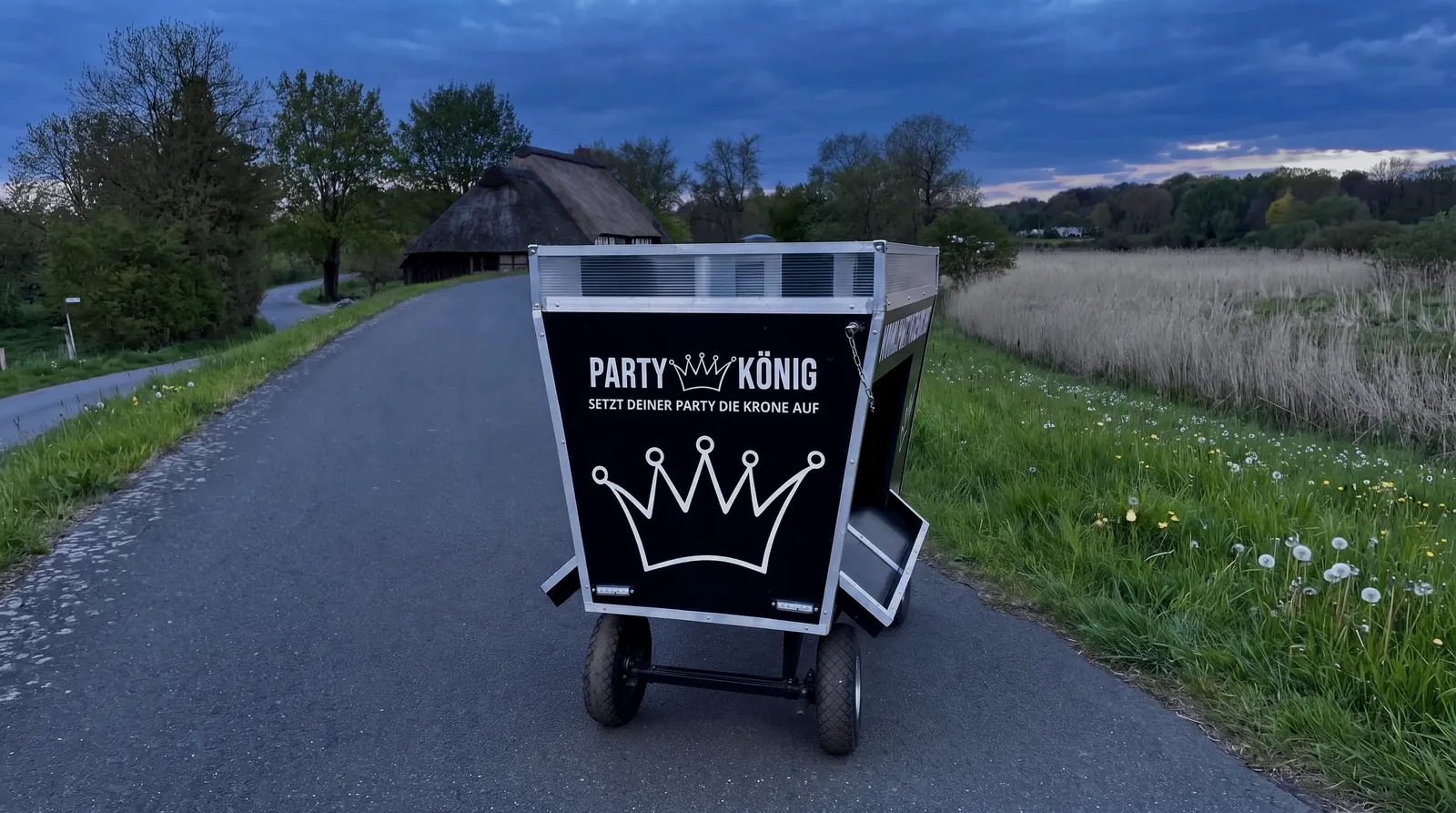 Der schwarze Partykönig-Bollerwagen von vorn, mit Logo und großer weißer Krone, in der blauen Stunde